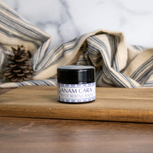 Jar of Anam Cara Restorative Balm on a wooden surface with a striped cloth behind it