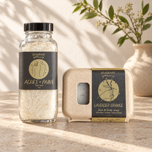 Seagrape bath soak and Lavender Orange soap on a countertop with a plant at the upper right; styling props not included.